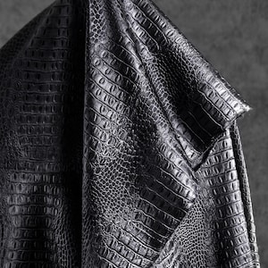 May include: Close-up of a black, textured fabric with a crocodile or alligator skin pattern. The material is folded, revealing the detailed scales and a subtle sheen. The fabric appears to be leather or a leather-like material.