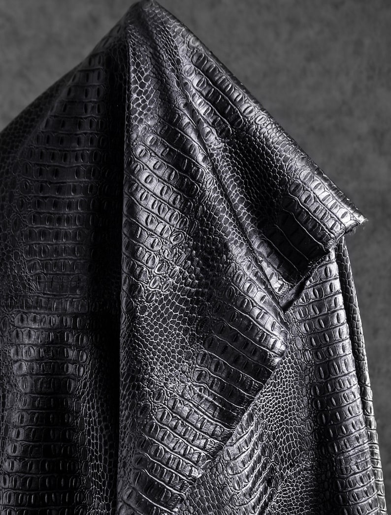May include: Close-up of a black, textured fabric with a crocodile or alligator skin pattern. The material is folded, revealing the detailed scales and a subtle sheen. The fabric appears to be leather or a leather-like material.