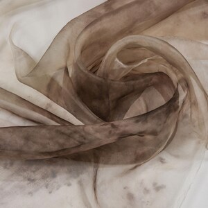 May include: A close-up of a sheer fabric with a swirling pattern. The fabric transitions from light beige to deep brown, with darker spots scattered throughout. The material appears delicate and flowing, with soft folds and curves.