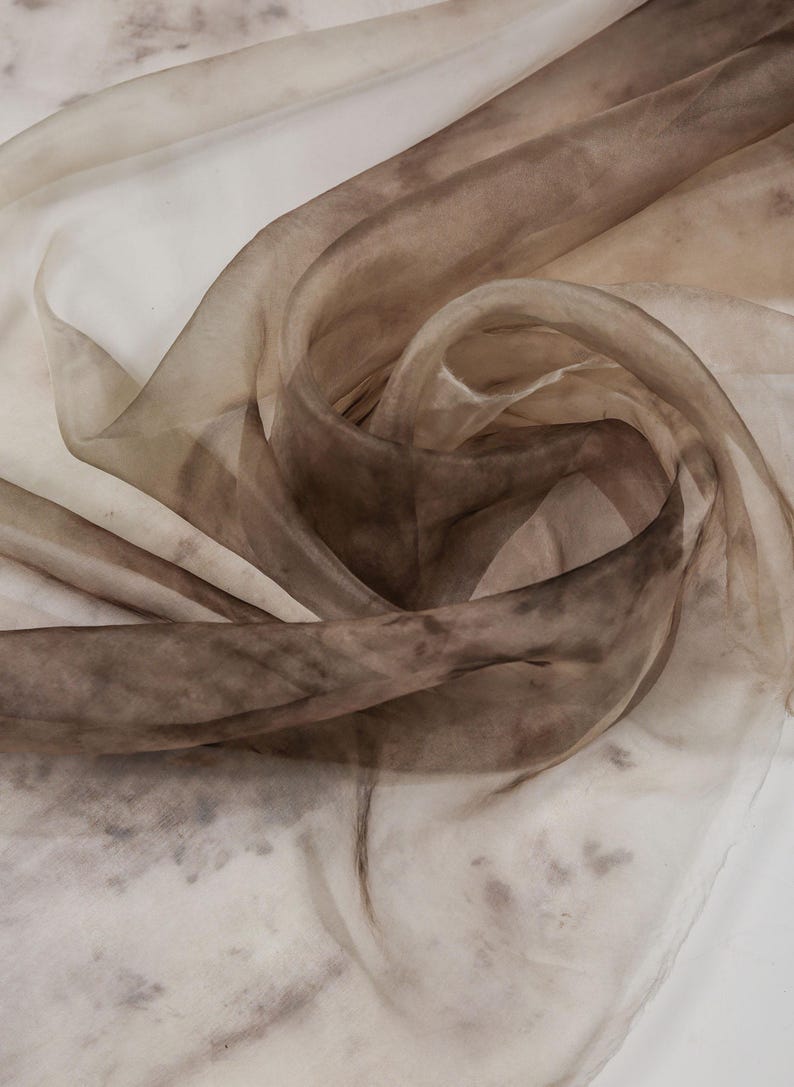 May include: A close-up of a sheer fabric with a swirling pattern. The fabric transitions from light beige to deep brown, with darker spots scattered throughout. The material appears delicate and flowing, with soft folds and curves.