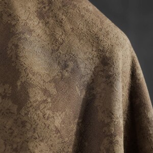 May include: Close-up of a textured fabric in shades of brown and tan. The fabric has a subtle, mottled pattern, creating a sense of depth and visual interest. The material appears to be draped, with soft folds and shadows.