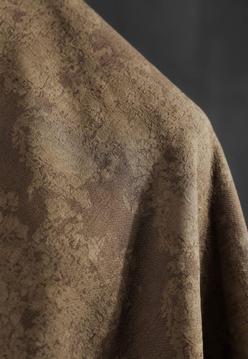 May include: Close-up of a textured fabric in shades of brown and tan. The fabric has a subtle, mottled pattern, creating a sense of depth and visual interest. The material appears to be draped, with soft folds and shadows.
