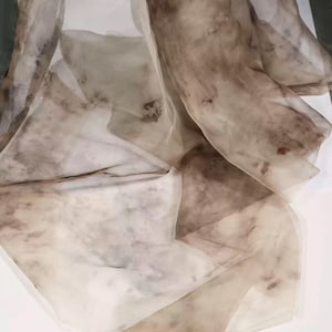 May include: A sheer, flowing fabric in shades of brown and beige. The material appears lightweight and delicate, with a marbled or watercolor-like pattern. The fabric is draped, creating soft folds and shadows. The overall effect is ethereal and artistic.