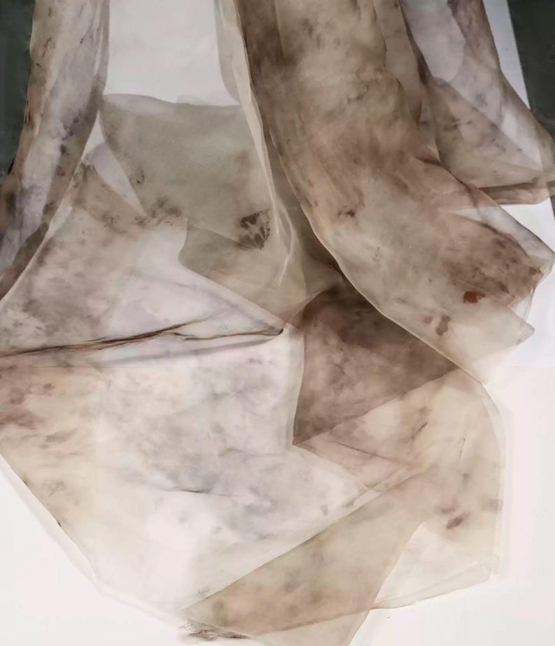 May include: A sheer, flowing fabric in shades of brown and beige. The material appears lightweight and delicate, with a marbled or watercolor-like pattern. The fabric is draped, creating soft folds and shadows. The overall effect is ethereal and artistic.