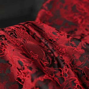K&ouml;nnte beinhalten: Nahaufnahme eines luxuri&ouml;sen Stoffes mit floralem Muster. Der Stoff ist tiefrot mit einem kontrastierenden schwarzen Hintergrund. Das komplizierte Design ist in den Falten des Materials sichtbar, was auf einen hochwertigen Textilstoff hindeutet.