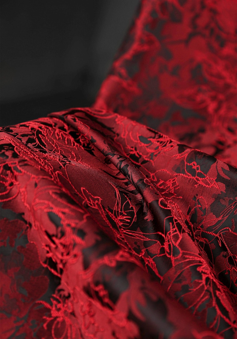 K&ouml;nnte beinhalten: Nahaufnahme eines luxuri&ouml;sen Stoffes mit floralem Muster. Der Stoff ist tiefrot mit einem kontrastierenden schwarzen Hintergrund. Das komplizierte Design ist in den Falten des Materials sichtbar, was auf einen hochwertigen Textilstoff hindeutet.