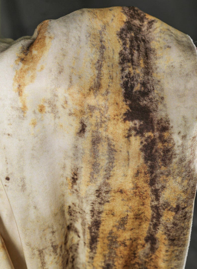 May include: Close-up of a fabric with a mottled pattern. The fabric has a cream-colored base with brown and tan streaks and blotches, creating a textured, organic appearance. The overall effect is reminiscent of animal hide.