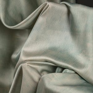 May include: Close-up of a flowing fabric in shades of pale green and gray. The material has a subtle sheen and a soft texture, with folds and drapes creating shadows. The overall effect is elegant and luxurious, suggesting a high-quality textile.