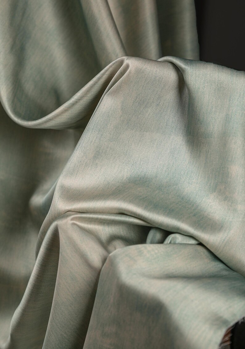May include: Close-up of a flowing fabric in shades of pale green and gray. The material has a subtle sheen and a soft texture, with folds and drapes creating shadows. The overall effect is elegant and luxurious, suggesting a high-quality textile.