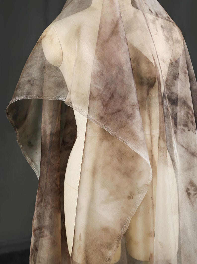 May include: A sheer, flowing fabric draped over a mannequin. The fabric features a mottled pattern in shades of brown and beige, creating a watercolor effect. The material appears lightweight and delicate, with visible seams.