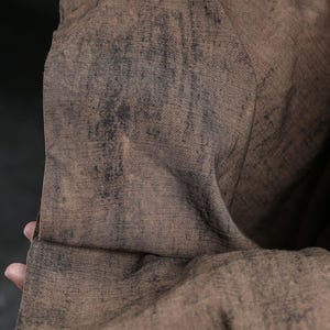 Puede incluir: Primer plano de una tela texturizada con un aspecto desgastado y moteado. La tela es principalmente marr&oacute;n con parches m&aacute;s oscuros, de color marr&oacute;n negruzco, creando una est&eacute;tica vintage o desgastada. El material parece suave y flexible.