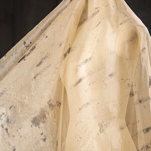 May include: A beige dress form is draped with a sheer, textured fabric. The fabric has dark gray and black mottled patterns. The dress form is in a studio setting, with a dark background.