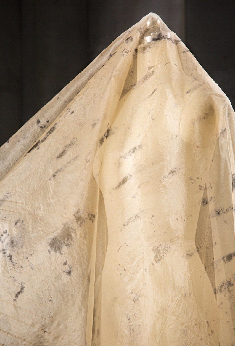 May include: A beige dress form is draped with a sheer, textured fabric. The fabric has dark gray and black mottled patterns. The dress form is in a studio setting, with a dark background.