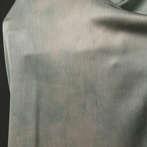 May include: A close-up of a flowing fabric in shades of pale blue and white. The material has a soft, textured appearance, with subtle variations in color creating a watercolor effect. The fabric is draped, showing its drape and texture.