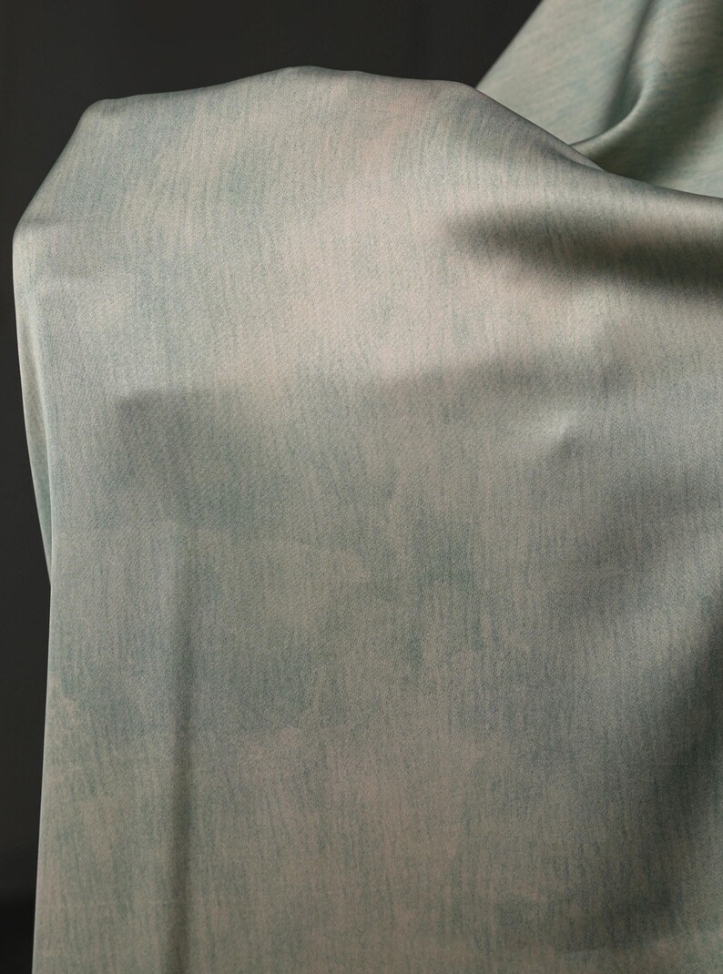 May include: A close-up of a flowing fabric in shades of pale blue and white. The material has a soft, textured appearance, with subtle variations in color creating a watercolor effect. The fabric is draped, showing its drape and texture.