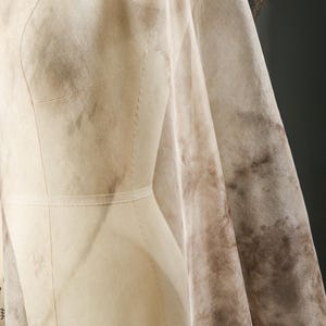 May include: A beige dress form draped with fabric. The fabric has a mottled brown pattern, creating a watercolor effect. The dress form is detailed with seams and a waistline, suggesting a garment in progress. The overall aesthetic is soft and textured.