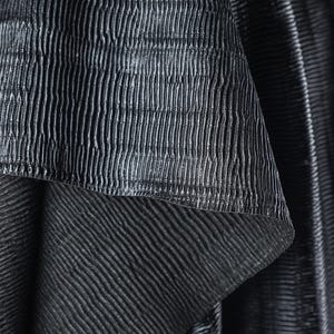 May include: Close-up of a textured, dark gray fabric with vertical and horizontal ribbed patterns. The fabric appears to be layered, with a top layer folded over to reveal a contrasting texture underneath. The overall effect is a play of light and shadow.