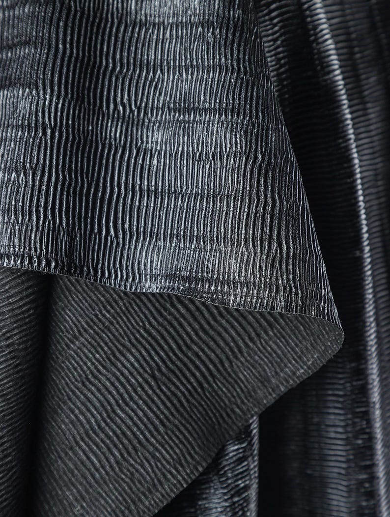 May include: Close-up of a textured, dark gray fabric with vertical and horizontal ribbed patterns. The fabric appears to be layered, with a top layer folded over to reveal a contrasting texture underneath. The overall effect is a play of light and shadow.