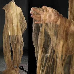 May include: A beige, semi-transparent fabric draped over a dress form. The fabric has a textured, wrinkled appearance and hangs in long, vertical strips. The dress form has a tag with the numbers "84" and "T" on it. The fabric is lit from the side, creating shadows.