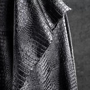 May include: Close-up of a textured, black fabric with a crocodile or alligator skin pattern. The material is folded, revealing the intricate details of the embossed design. The fabric has a slight sheen, suggesting a leather or leather-like material.