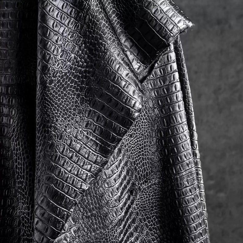 May include: Close-up of a textured, black fabric with a crocodile or alligator skin pattern. The material is folded, revealing the intricate details of the embossed design. The fabric has a slight sheen, suggesting a leather or leather-like material.