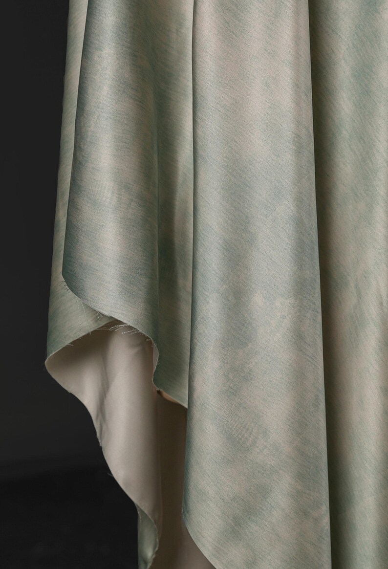 May include: Close-up of draped fabric with a watercolor effect. The fabric has a light green and blue color palette with a subtle texture. The underside of the fabric is a light beige color.