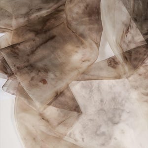 May include: Close-up of flowing, sheer fabric in shades of brown and beige. The fabric appears to be layered and has a marbled or watercolor effect, with darker brown patches and subtle variations in tone. The overall effect is soft and ethereal.
