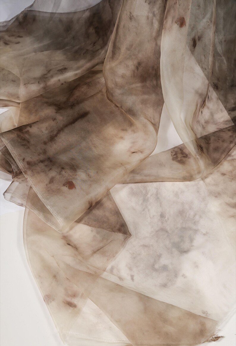 May include: Close-up of flowing, sheer fabric in shades of brown and beige. The fabric appears to be layered and has a marbled or watercolor effect, with darker brown patches and subtle variations in tone. The overall effect is soft and ethereal.