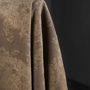 May include: Close-up of a draped fabric in shades of brown and tan. The textile has a textured, mottled pattern, with areas of lighter and darker tones creating a visual depth. The fabric appears to be thick and possibly suitable for upholstery or drapery.