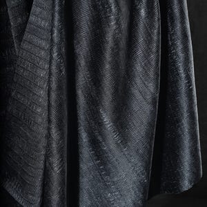 May include: Close-up of a dark, textured fabric with a subtle sheen. The material is draped, creating folds and shadows that highlight the diagonal ribbed pattern. The overall effect is a luxurious, elegant textile.