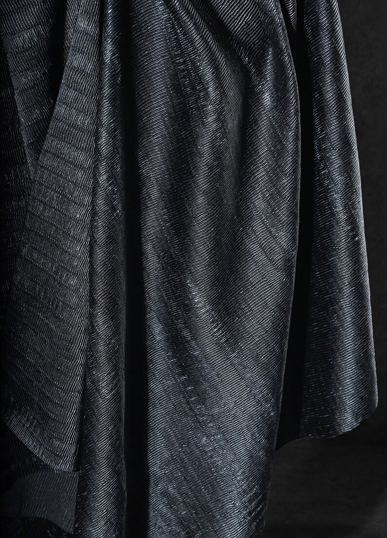 May include: Close-up of a dark, textured fabric with a subtle sheen. The material is draped, creating folds and shadows that highlight the diagonal ribbed pattern. The overall effect is a luxurious, elegant textile.