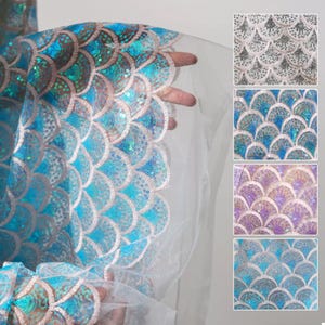 Può includere: Primo piano di un tessuto trasparente con un motivo a scaglie di sirena. Il tessuto è azzurro con paillettes iridescenti e un retro bianco. Vengono mostrati campioni di tessuto aggiuntivi in argento, blu e viola.
