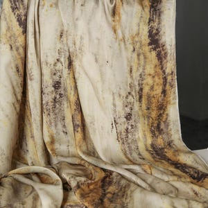 May include: A draped fabric with a marbled pattern in shades of cream, brown, and gold. The fabric is soft and flowing, with vertical streaks and a textured appearance. The overall effect is reminiscent of natural stone or wood grain.