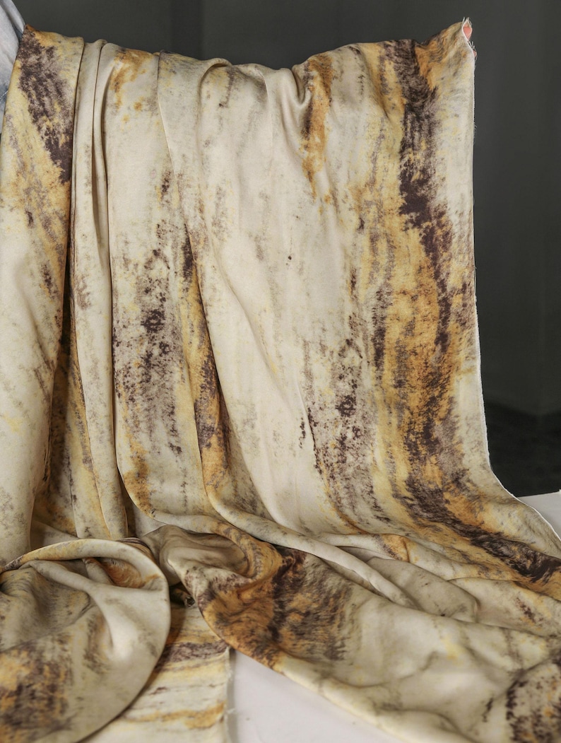 May include: A draped fabric with a marbled pattern in shades of cream, brown, and gold. The fabric is soft and flowing, with vertical streaks and a textured appearance. The overall effect is reminiscent of natural stone or wood grain.