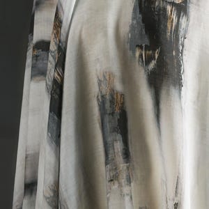 May include: Close-up of a fabric with an abstract design. The fabric is mainly off-white with vertical brushstrokes in black, grey, and touches of gold. The fabric is draped, showing its texture and flow.