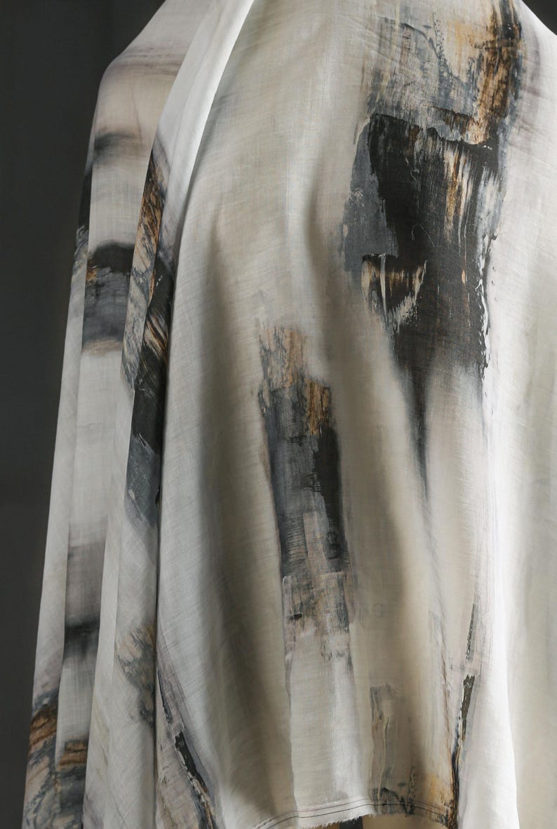 May include: Close-up of a fabric with an abstract design. The fabric is mainly off-white with vertical brushstrokes in black, grey, and touches of gold. The fabric is draped, showing its texture and flow.