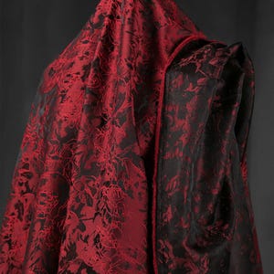 K&ouml;nnte beinhalten: Ein Kapuzenmantel aus rot-schwarz gemustertem Stoff. Die Au&szlig;enseite des Umhangs ist tiefrot mit einem detaillierten floralen Design, w&auml;hrend die Innenseite und die Verzierungen schwarz sind. Der Stoff scheint luxuri&ouml;s zu sein.