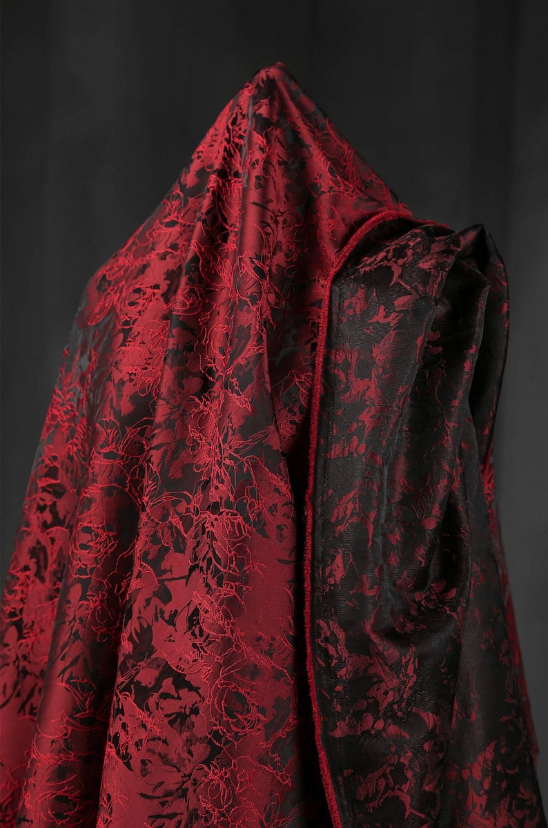 K&ouml;nnte beinhalten: Ein Kapuzenmantel aus rot-schwarz gemustertem Stoff. Die Au&szlig;enseite des Umhangs ist tiefrot mit einem detaillierten floralen Design, w&auml;hrend die Innenseite und die Verzierungen schwarz sind. Der Stoff scheint luxuri&ouml;s zu sein.