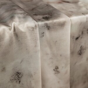 May include: Close-up of a folded, light-colored fabric with a marbled pattern. The fabric has a soft, flowing appearance with dark gray and black splotches and a few red accents. The folds create shadows and depth.
