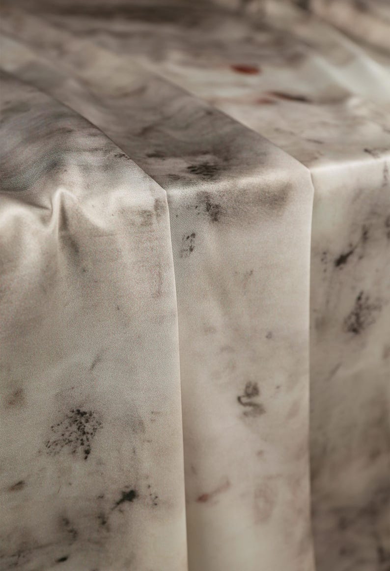 May include: Close-up of a folded, light-colored fabric with a marbled pattern. The fabric has a soft, flowing appearance with dark gray and black splotches and a few red accents. The folds create shadows and depth.