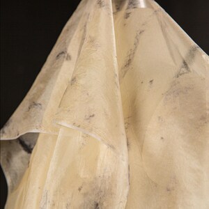 May include: A draped, semi-transparent fabric with a pale yellow hue and subtle dark gray mottled patterns. The fabric is folded, creating soft shadows and highlights against a black background. The texture appears slightly crinkled.