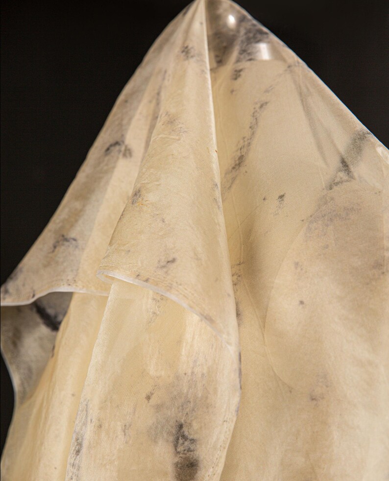 May include: A draped, semi-transparent fabric with a pale yellow hue and subtle dark gray mottled patterns. The fabric is folded, creating soft shadows and highlights against a black background. The texture appears slightly crinkled.