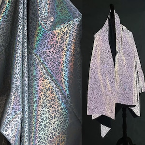 Op de afbeelding: Zilverkleurige stof met een geometrisch patroon en een bijpassende jas. De stof heeft een reflecterende, iriserende kwaliteit, met een netwerk van lijnen dat een textuur creëert. De jas is gedrapeerd op een zwarte mannequin.