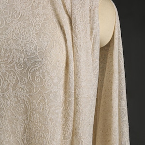 May include: Ivory-colored fabric with an intricate floral pattern, draped over a mannequin. The fabric has a delicate, textured appearance, with detailed swirling designs. The material is soft and flowing, suggesting it could be used for clothing or home decor.