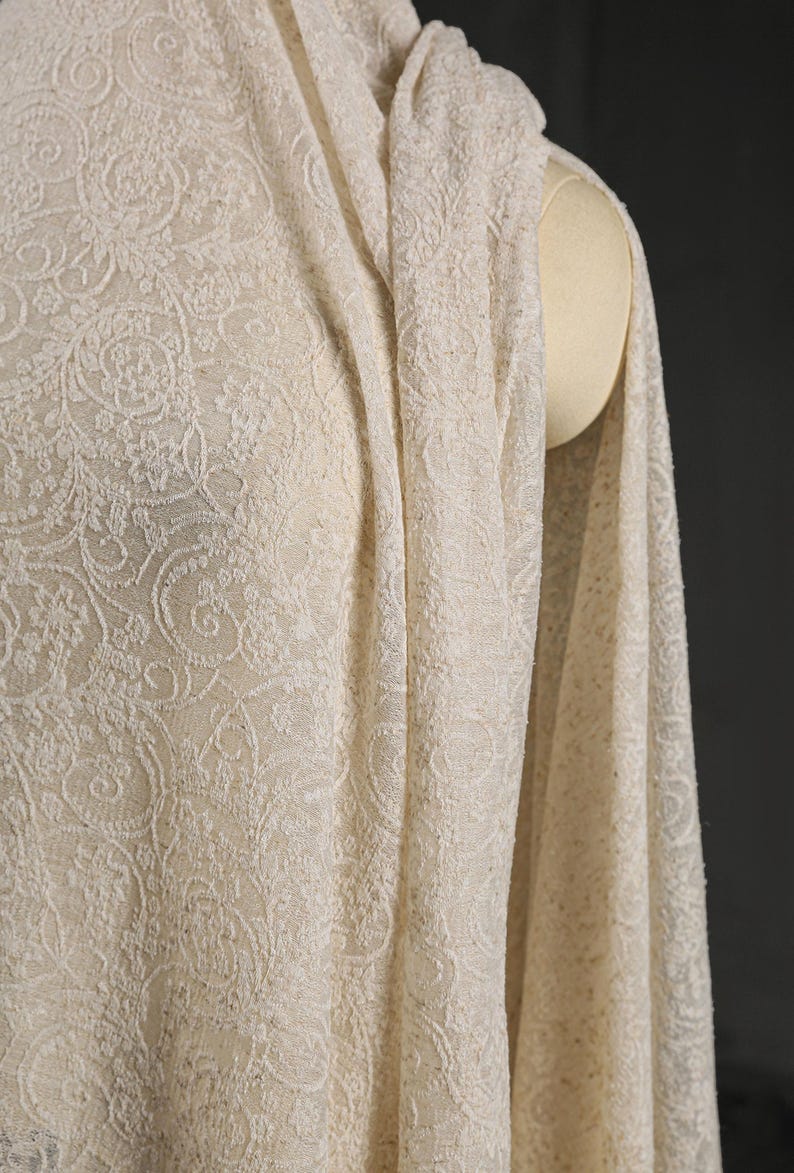 May include: Ivory-colored fabric with an intricate floral pattern, draped over a mannequin. The fabric has a delicate, textured appearance, with detailed swirling designs. The material is soft and flowing, suggesting it could be used for clothing or home decor.