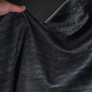 May include: Close-up of a dark gray fabric with a textured, ribbed pattern. The fabric is draped, creating shadows and highlights that emphasize the texture. The material appears to be soft and flowing.