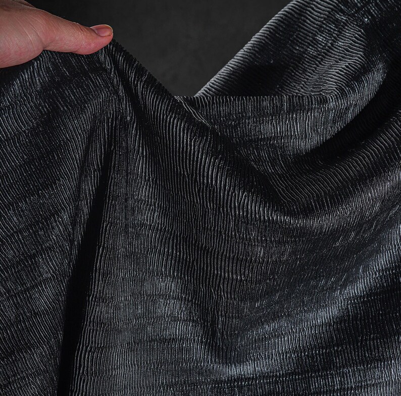 May include: Close-up of a dark gray fabric with a textured, ribbed pattern. The fabric is draped, creating shadows and highlights that emphasize the texture. The material appears to be soft and flowing.