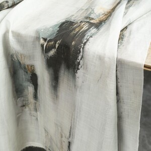 May include: A white tablecloth with an abstract design. The design features dark grey, black, and brown brushstrokes, creating a textured, artistic look. The fabric drapes over a wooden table, showcasing the pattern.