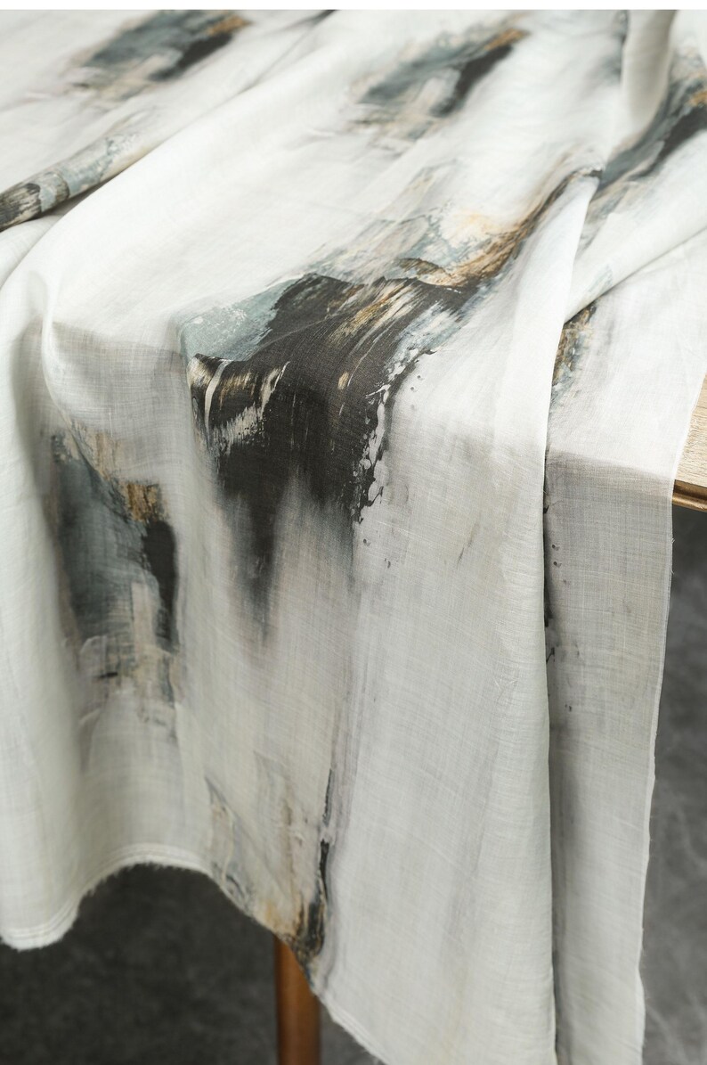 May include: A white tablecloth with an abstract design. The design features dark grey, black, and brown brushstrokes, creating a textured, artistic look. The fabric drapes over a wooden table, showcasing the pattern.