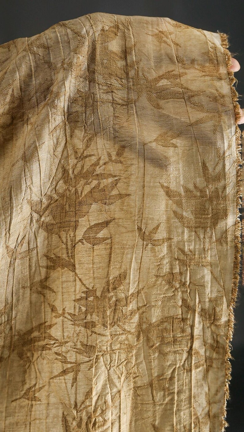 May include: Close-up of a beige fabric with a repeating pattern of brown leaves and stems. The fabric has a textured, pleated appearance and a frayed edge on the right side. The overall aesthetic is natural and organic.